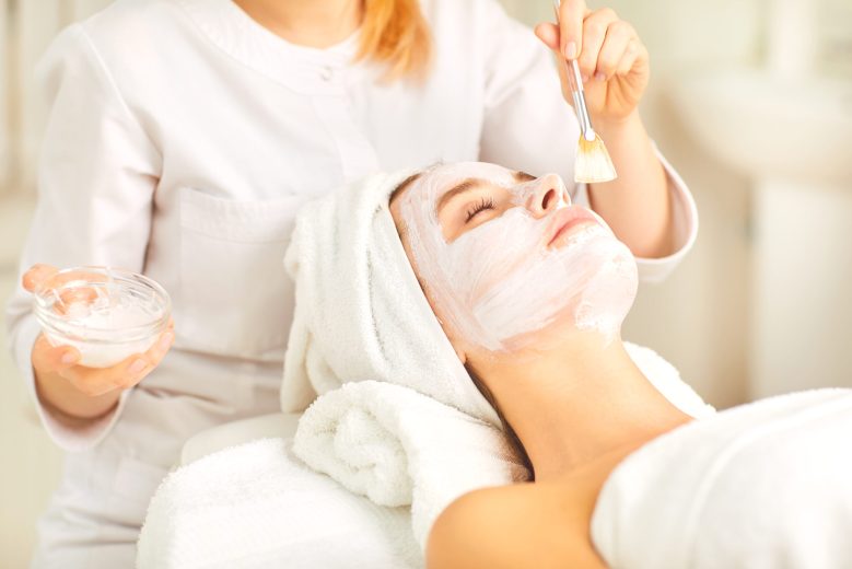 Beautician applies a white mask to a woman in the spa salon.